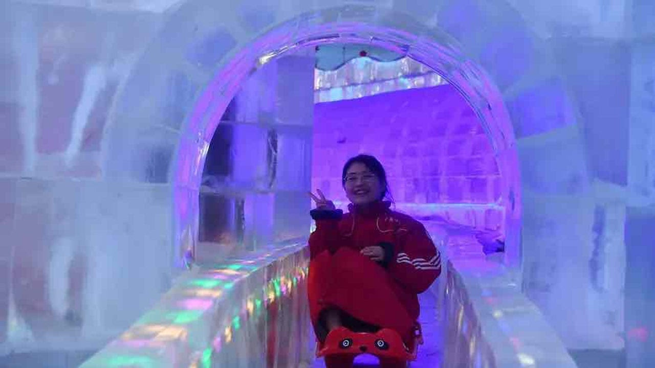 A smiling visitor poses in the ice sculpture hall. There are colorful lights inside the ice.