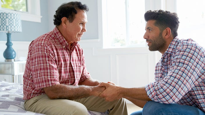 An adult son holds his father's hands while sitting down and talking to him at home.