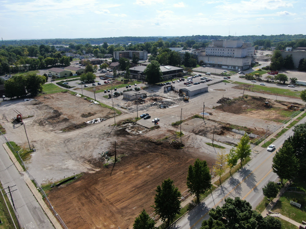 photo is looking southwest from the intersection of 2nd and Morton Street, there is a cleared site with exposed earth and the same equipment as the left photo working in the middle ground, the legacy hospital is shown in the background.