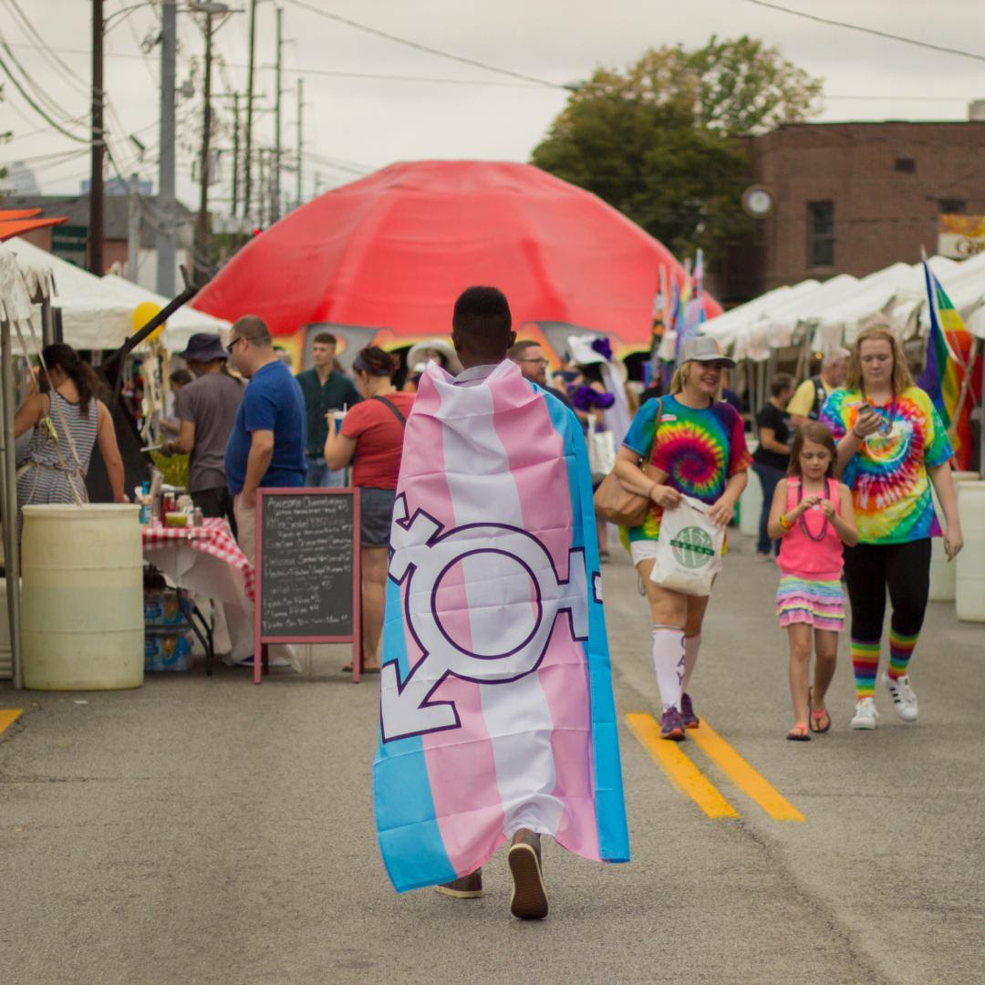 Photo of a person at a Pride event with their back to the camera. They are in front of a red umbrella and wrapped in a Trans Pride flag. 