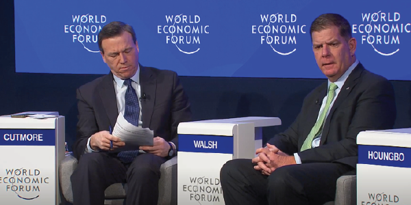 Secretary Walsh speaks as part of a panel. He is seated between a man in a suit, whose nameplate reads ‘Cutmore,’ and an unseen man whose nameplate reads ‘Houngbo.’ World Economic Forum icons cover the wall behind them.
