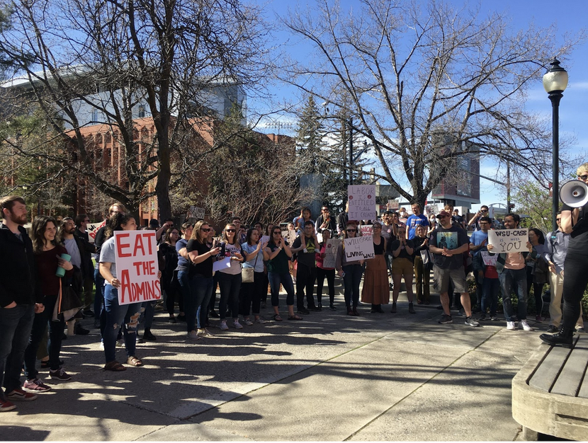 a group of people standing outside, looking at a speaker holding a megaphone. There are signs held up, including one that says "Eat the Admins"