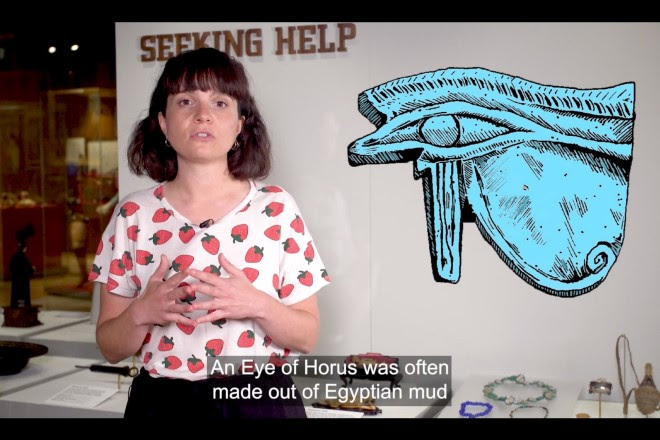 Screenshot of a YouTube video, featuring a person in a shirt with red strawberries on it, standing in front of the Medicine Man galleries in Wellcome Collection, with an illustration of the eye of horus superimposed on top, and the caption "An Eye of Horus was often made out of Egyptian mud" at the bottom