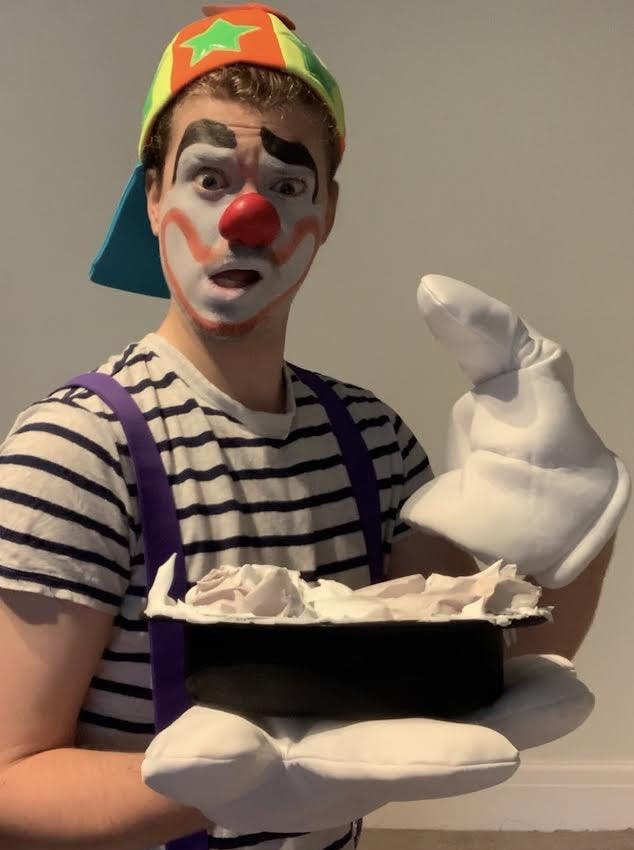 Barney in clown kink gear, holding a fake pie while wearing very large white gloves.