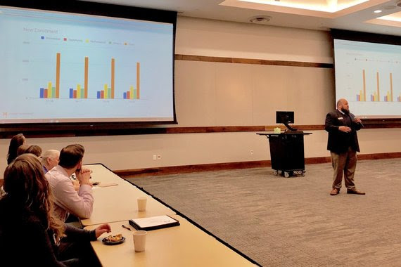 WDE Hathaway consultant, Bradley Barker, speaks to the Leadership Cheyenne class in a classroom at Laramie County Community College, with graphs projected on screens behind him.