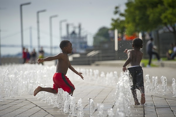 Two little boys, one in navy blue swim trunks, the other in red, chase each other on a concrete water splash pad set in an urban walkway