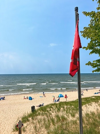 A red beach warning flag in the foreground, with people and umbrellas dotting the sandy beach behind. It's a sunny, blue sky day.