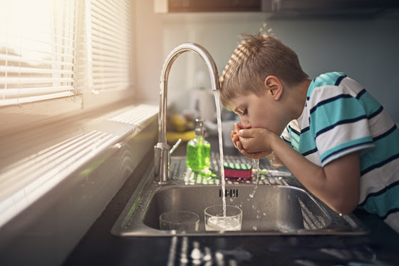 Boy drinking water from a kitchen sink faucet while in front of window