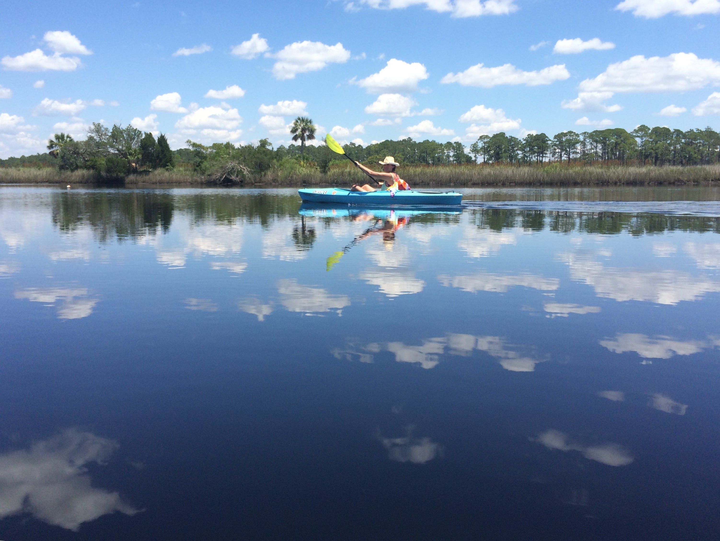 Woman paddles through water so calm it is reflecting the clouds and sky.