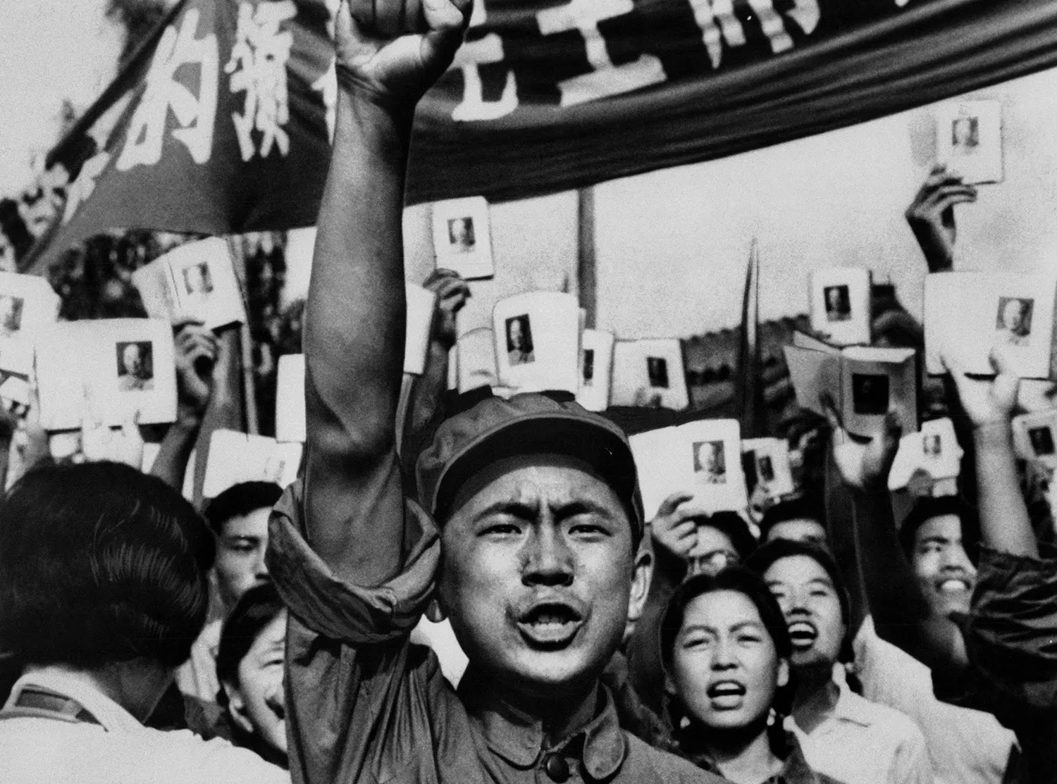 A soldier holds up his fist as a crowd of people behind him cheer holding up images in books and on pieces of paper under a banner with Chinese characters on it.