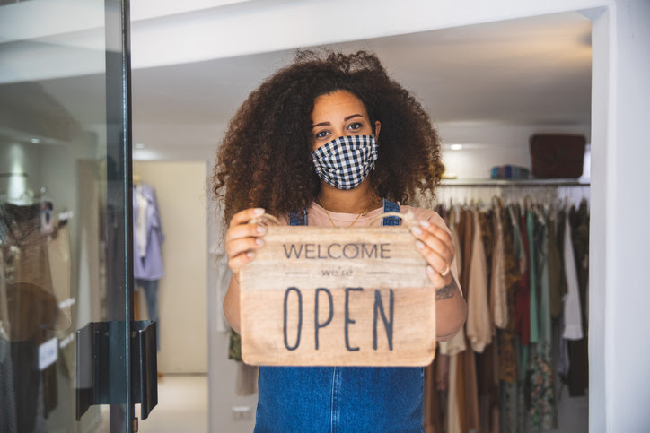 A retail worker wearing a face mask holds up a sign to indicate that her clothing store is open during the COVID-19 pandemic.