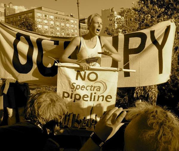A woman hold a stop Spectra sign in front of an Occupy banner while giving a speech to a crowd