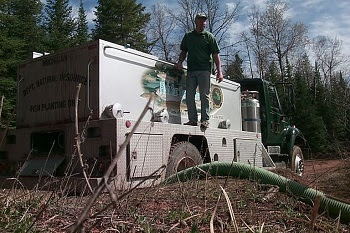 A man dressed in green T-shirt and khakis stands on DNR fish stocking truck, overseeing distribution of fish through a large green hose