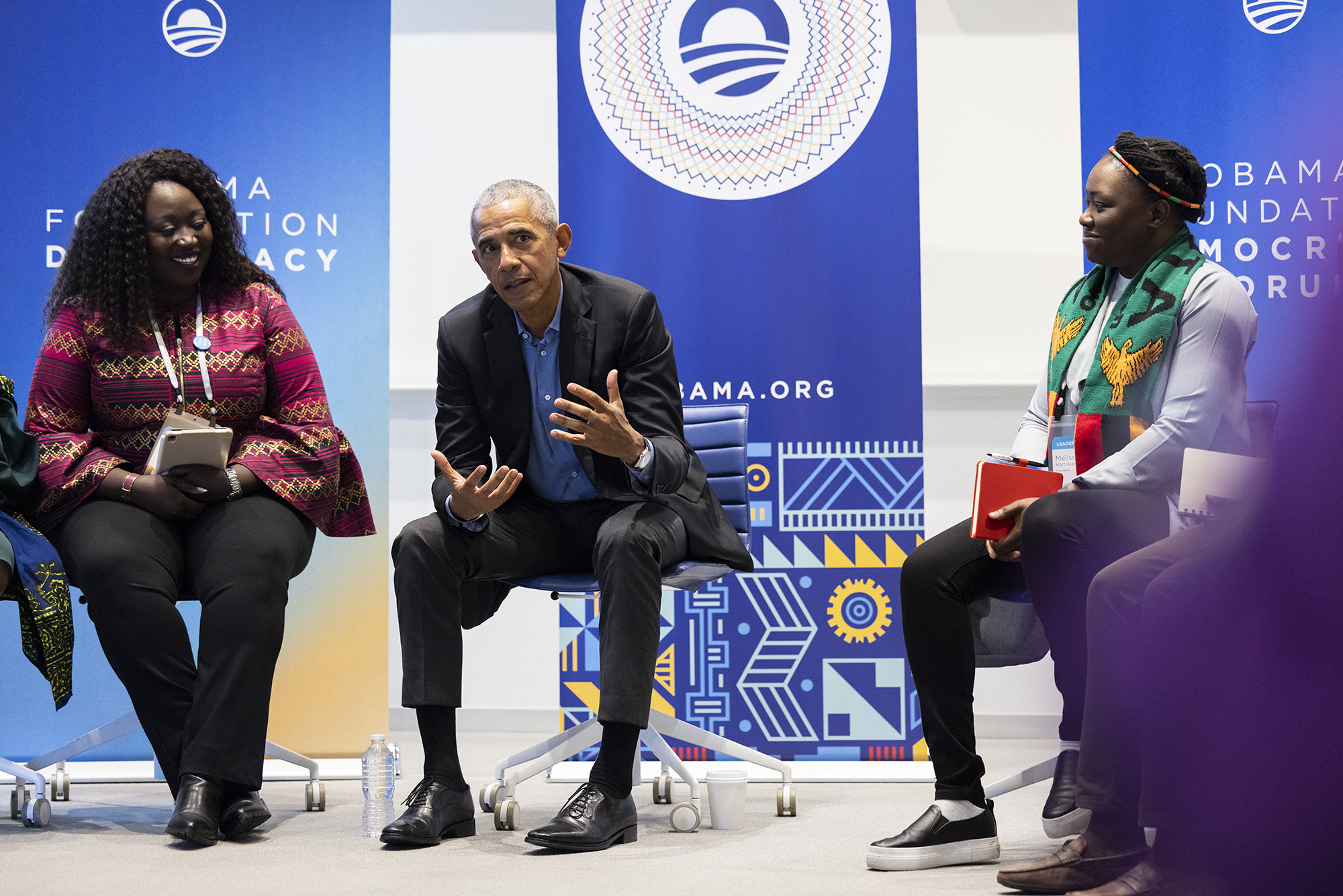 President Obama sits on a stage between a young women and a young man with deep skin tones. In the background are blue and white graphics that read, “Obama.org” and “The Obama Foundation Democracy Forum.” 