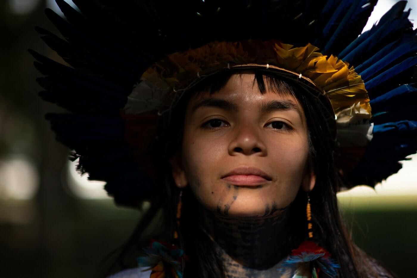 Txai é uma jovem indígena com cabelos e olhos castanhos. Ela usa um cocar na cabeça, brinco de pena nas orelhas e tem pintura étnica no rosto e pescoço