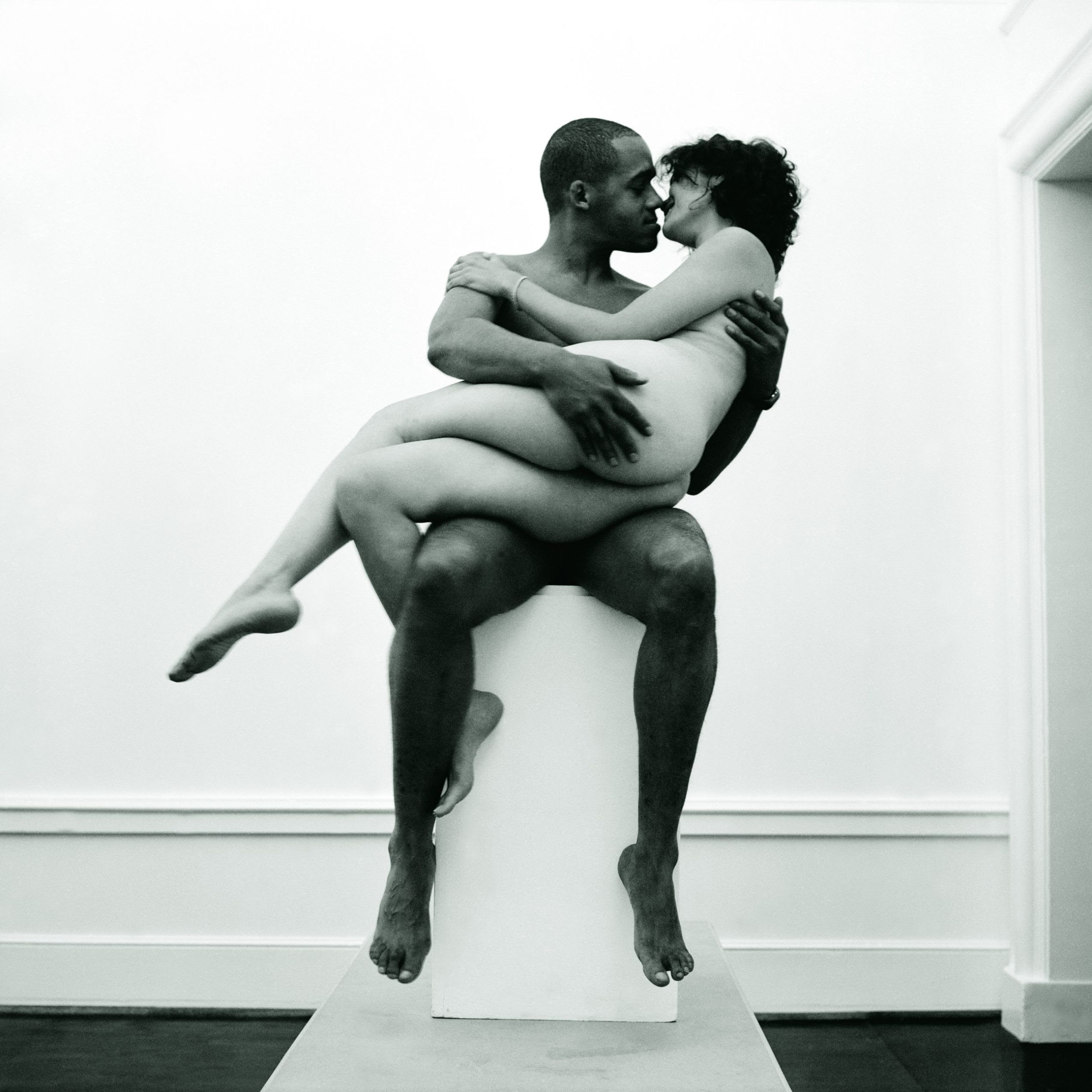 A photographic work by Tracey Rose. The black and white image features a brown-skinned man and a light-skinned woman embracing, naked, while sitting on a white pedestal. Posing against a white wall, smiling, their noses are touching. They seem to be on the verge of kissing. 