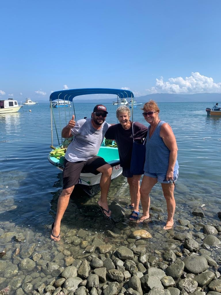 Three adults stand in shallow water in front of a small boat, smiling at camera