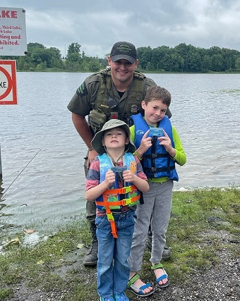 a man in a DNR conservation officer uniform with a young boy and girl wearing life jackets stand on the grassy shore of a lake