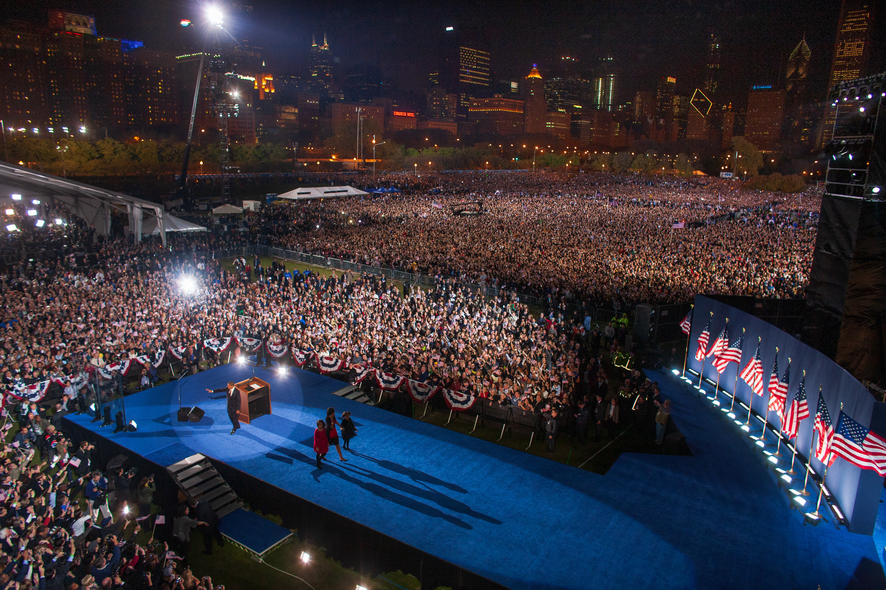 An aerial shot of the crowd at Grant Park on Election Night 2008 with President Obama at the podium waving at the crowd and Mrs. Obama walking off the stage with Sasha and Malia Obama.
