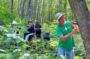 Man in green T-shirt, shorts and tan, brimmed hat uses an axe to scrape bark on an oak tree, while a group of young men and women watch.