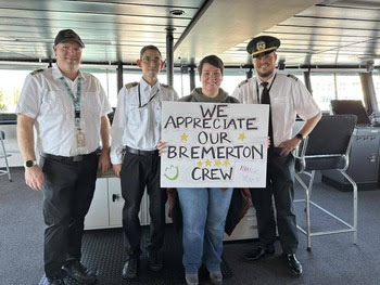 Four people in the wheelhouse of a ferry, three crewmembers and the other holding a "We appreciate our Bremerton crew" poster 