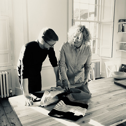 A photograph of makers Alan Dibble and Emma Green standing up at a table looking at a textiles piece. The photograph is in black and white with sepia tons.