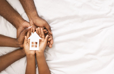 Family holds hands together with a small paper house in their palms.