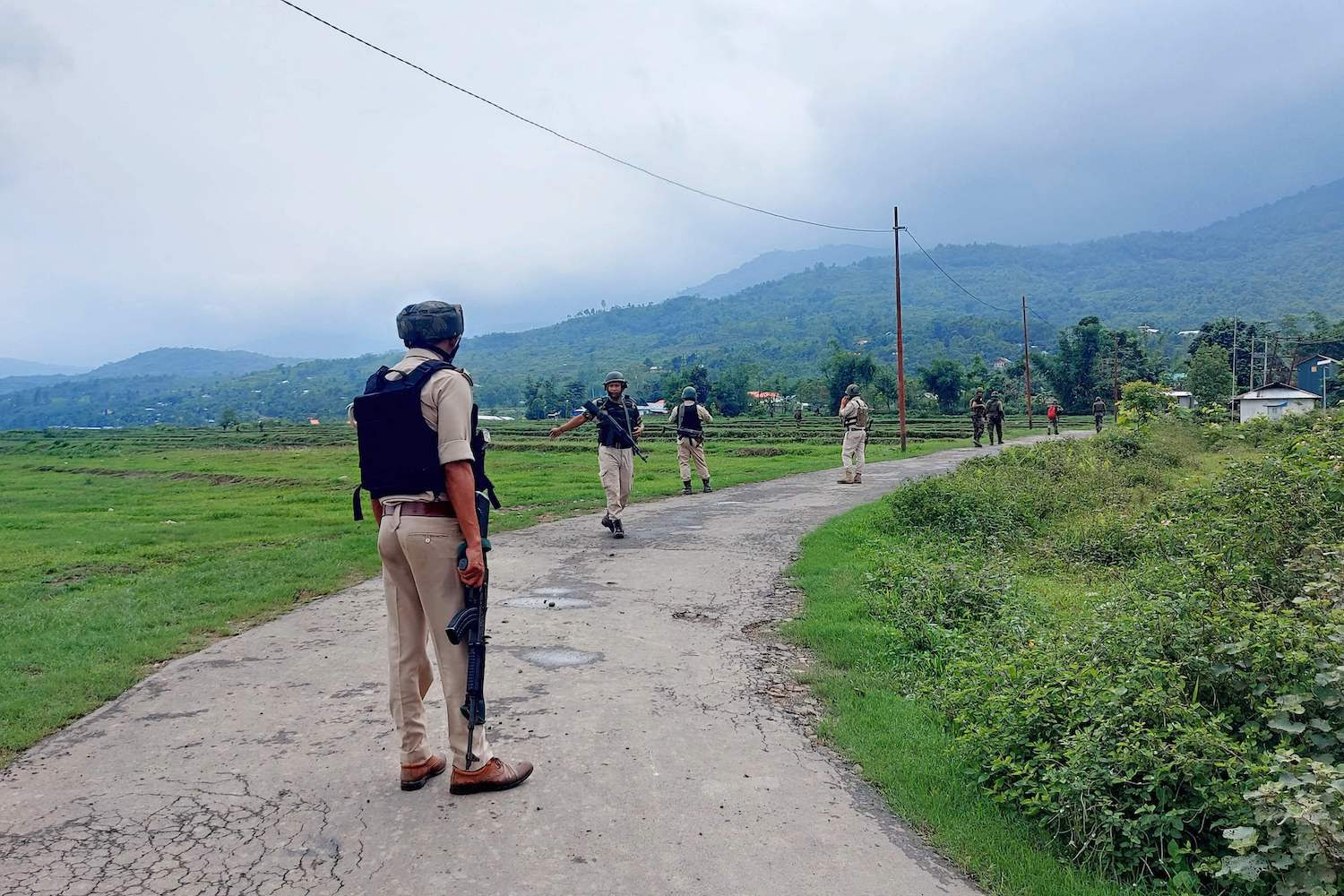 Uniformed Indian Army and police officers walk along a curved road in a foggy green valley. Some of the officers carry rifles, and most wear helmets.