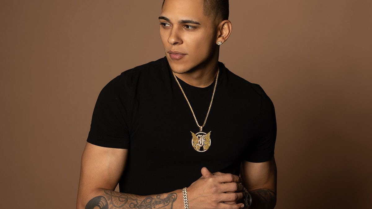 A young man wearing a black t-shirt and gold necklace shows tattoos on his forearms