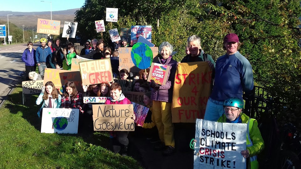 A group of around 30 strikers of all ages, holding signs in front of a large green hedge.