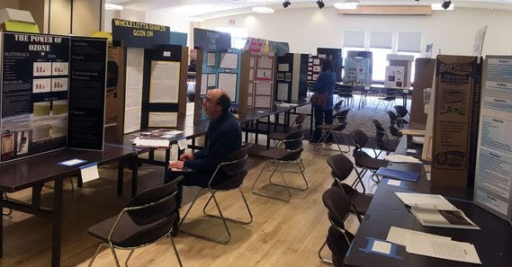 Rows of tables are filled with science project displays. A man sits and reads about the project titled "Whole Lotta Shakin Going On" which is next to a display titled "The Power of Ozone."
