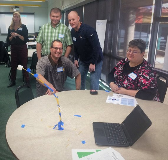 A group of teachers sit around a table observing an experiment with a marshmallow on pencils taped together.