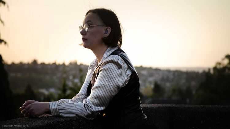 A  white woman in profile with short hair wearing purple hearing aids,  leather vest, pearl necklace, and striped button-down top, looking into  distance with Seattle skyline behind her.