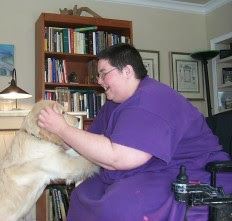 Carrie (purple dress; short hair; wheelchair) playing with a young golden retriever who has his paws on her knees.