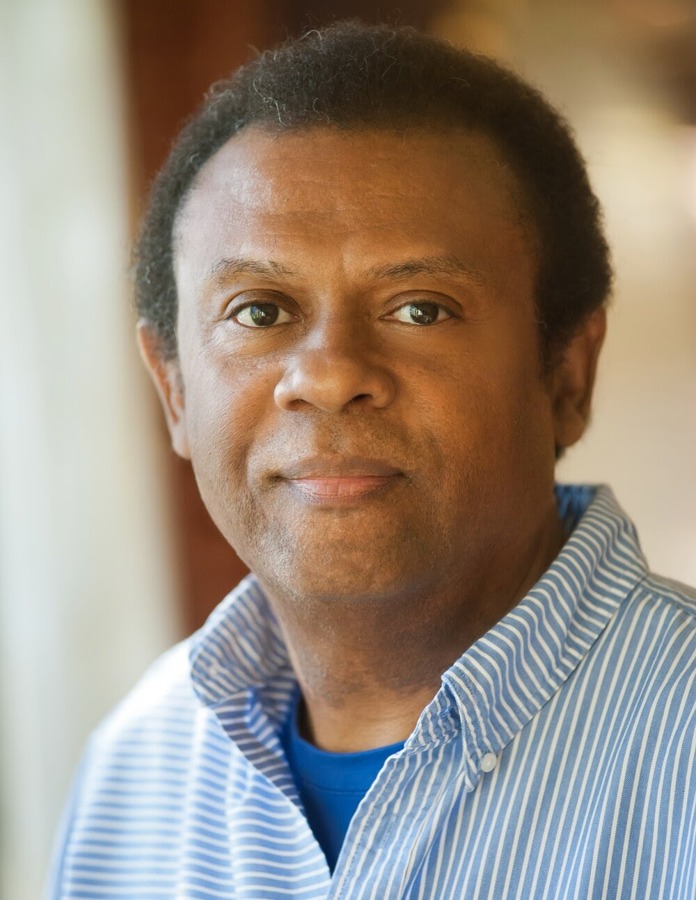 Headshot of Dr. Nicky Sheats, Black male wearing a blue shirt and blue and white striped collared shirt. Blurred tan and brown background.