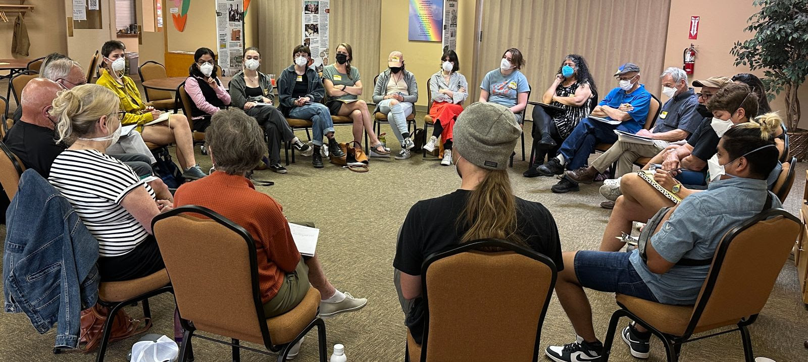 Around 20 people sit in a circle with masks on, mid-discussion.
