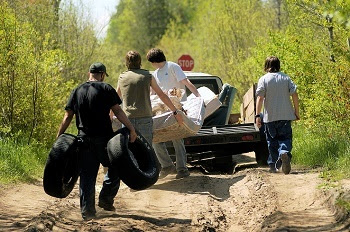 Four men in jeans and T-shirts carry old tires, a mattress and other garbage to a pickup truck parked on a dry, rutted, dirt road