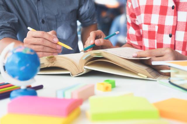 two students_ hands holding pencils to write in a book at a table