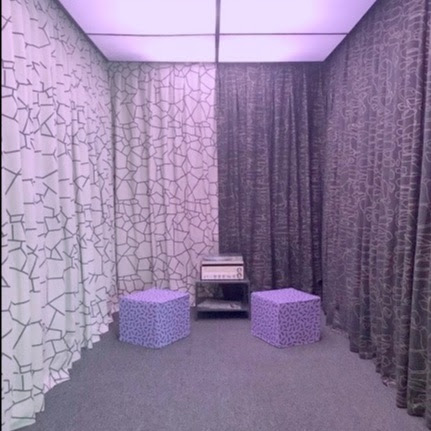 Photograph of the inside of an elevator with gray carpet, white and charcoal colored curtains, two purple cubes for people to sit on, and a record player.